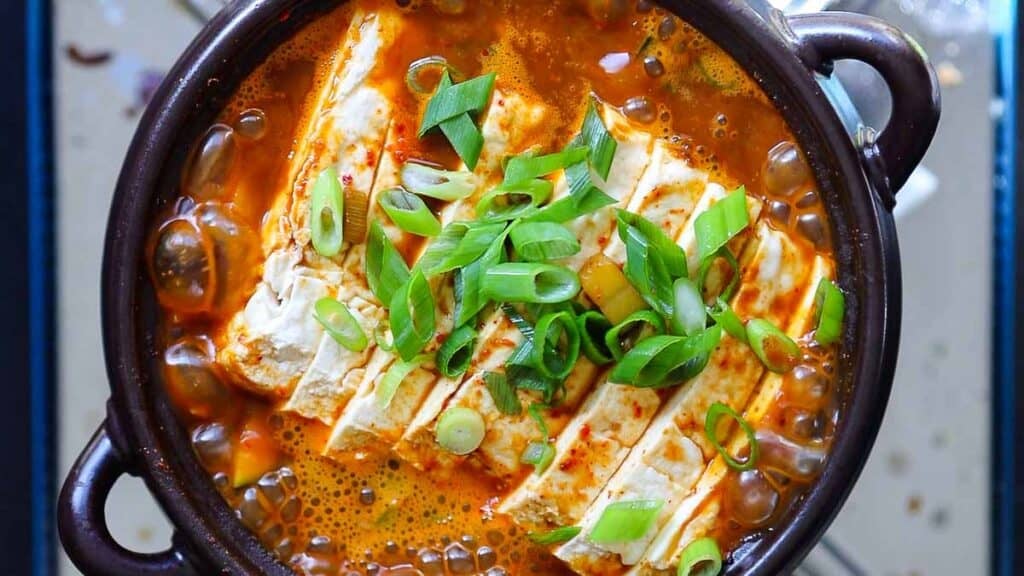 A bowl of spicy doenjang jjigae with a rich orange broth, topped with sliced green onions. The tofu is cut into rectangular pieces and arranged neatly in the soup, which is served in a dark ceramic bowl.